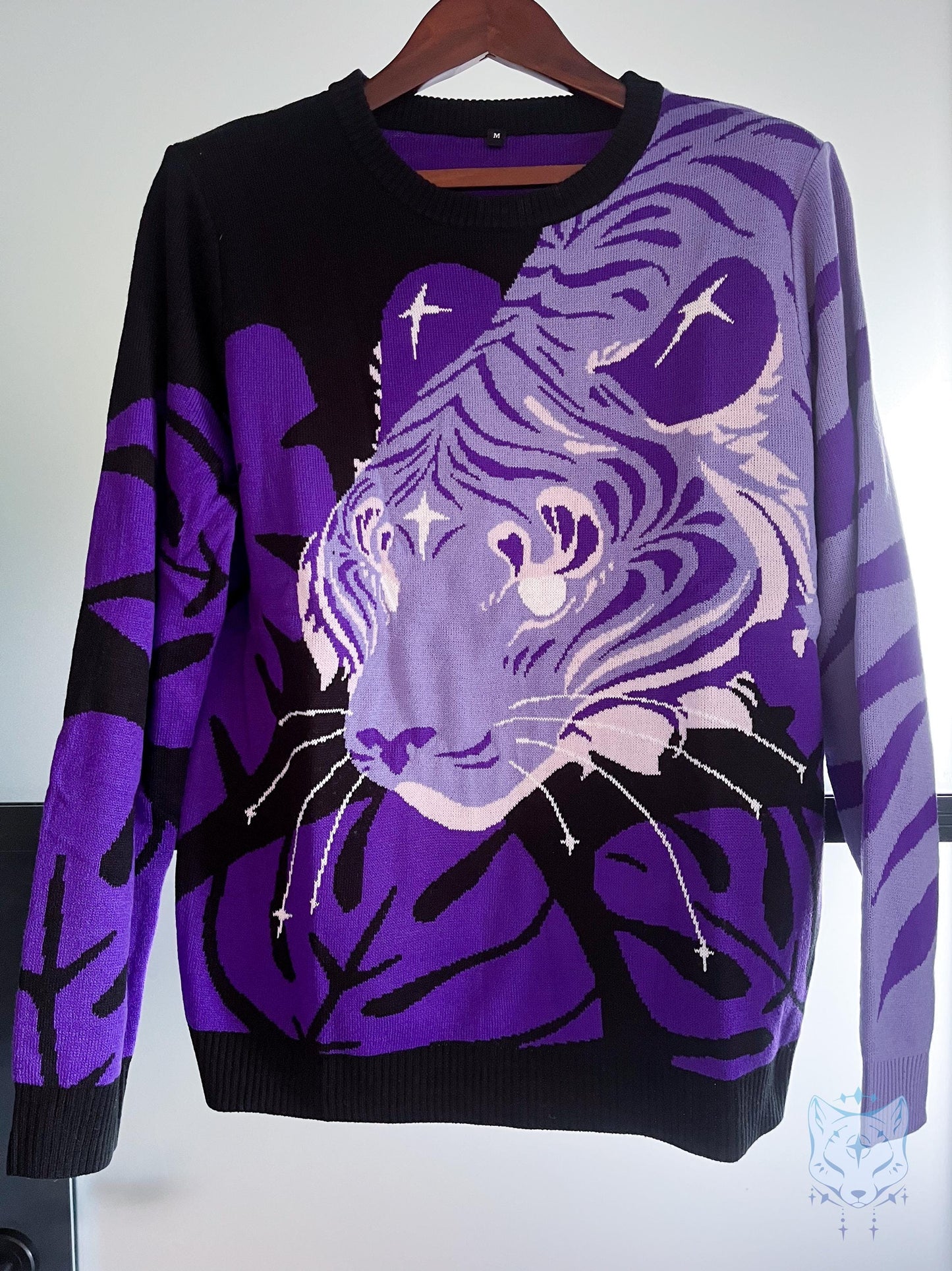 May include: A black and purple jumper with a tiger face design. The jumper has a crew neck and long sleeves. The tiger's face is a gradient of purple and white, with black accents. The jumper is hanging on a wooden hanger.