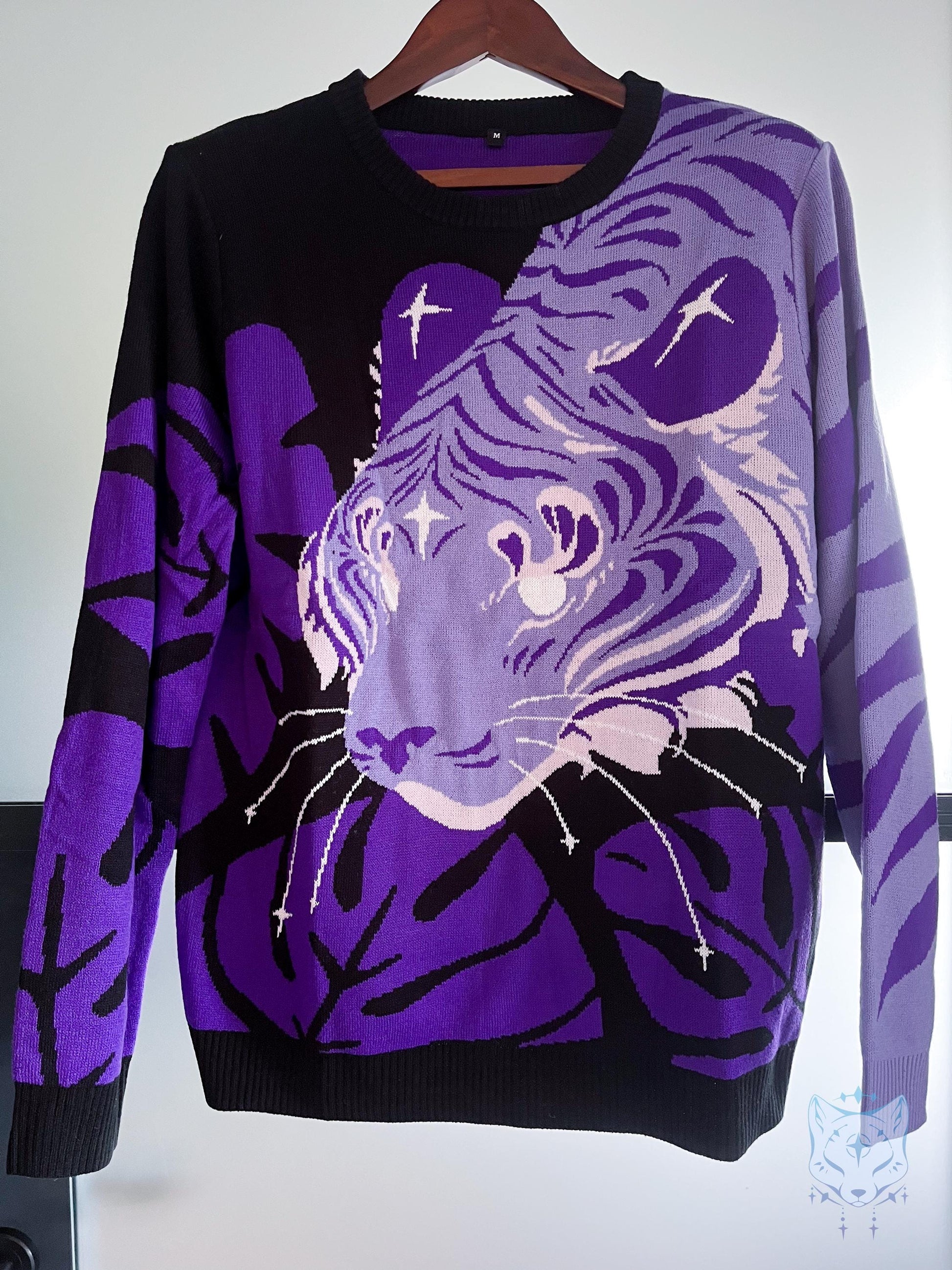 May include: A black and purple jumper with a tiger face design. The jumper has a crew neck and long sleeves. The tiger's face is a gradient of purple and white, with black accents. The jumper is hanging on a wooden hanger.
